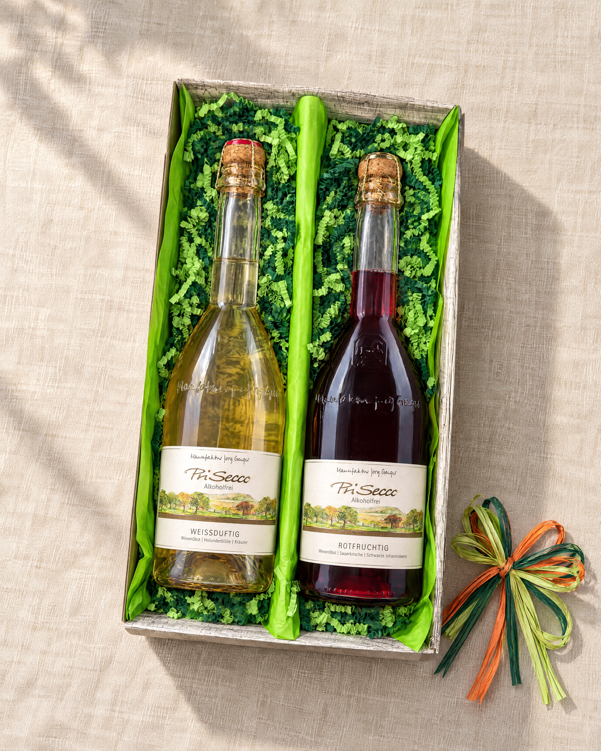 PriSecco Geschenkset mit Weißduftig und Rotfruchtig im offenen Zweierkarton, hellgrünem Papier, grüner Papierwolle und dreifarbiger Bastschleife